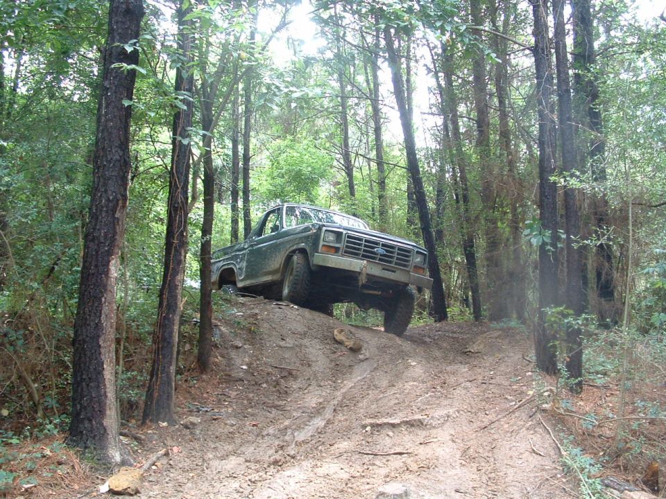 Green Ford F-Series truck climbing a muddy hill in a forest. Grille indicates 1980s model
