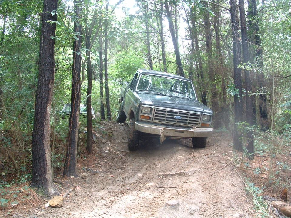 Ford Bronco driving on a muddy trail in the woods. Another vehicle is visible behind it