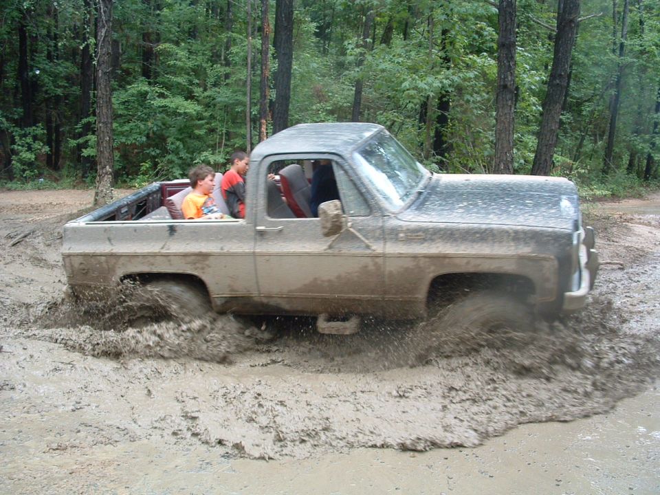 Old pickup truck with two kids in the cab driving through deep mud. The truck appears to be from the mid-1970s