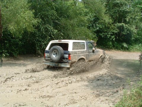 1993 Ford Bronco - A muddy 1993 Ford Bronco navigates a challenging off-road course, kicking up dirt and water.