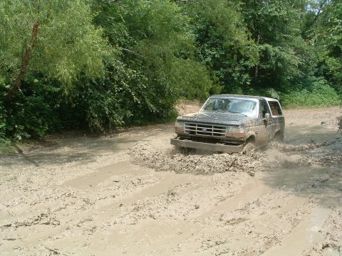 1993 Ford Bronco - A Ford Bronco navigates a muddy trail, kicking up water and dirt.