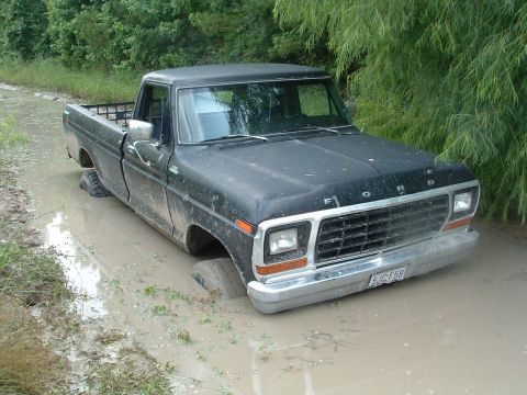 1993 Ford Bronco - A black Ford pickup truck is stuck in a muddy ditch, with trees and foliage in the background.