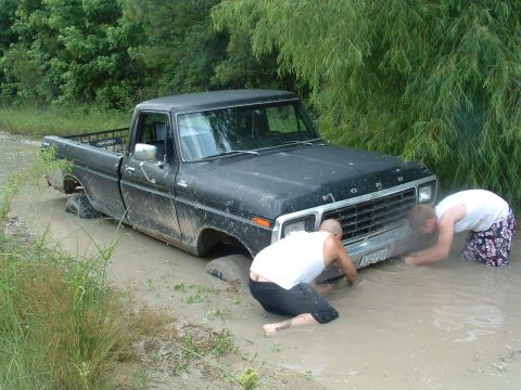 1993 Ford Bronco - Two people attempt to free a black 1979 Ford F-Series pickup truck that is stuck deep in mud.