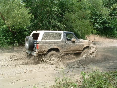 1993 Ford Bronco - A muddy Ford Bronco navigates a challenging off-road course, kicking up significant spray.