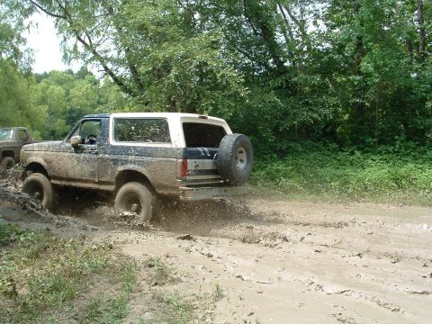 1993 Ford Bronco - A muddy 1993 Ford Bronco navigates a challenging off-road trail, kicking up mud as it goes.