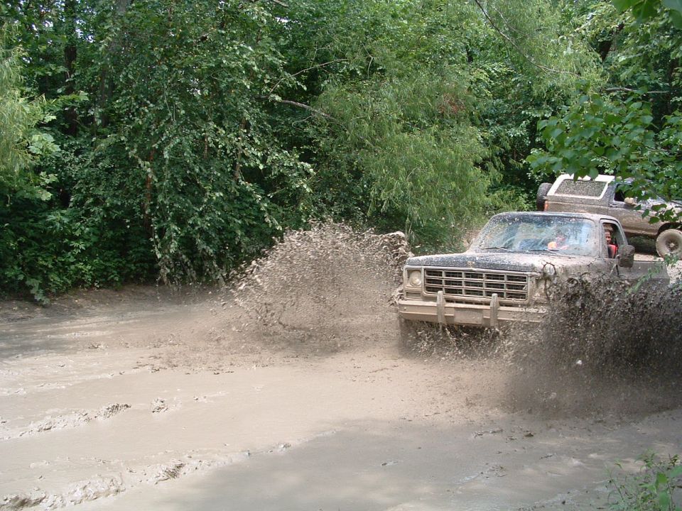 Two trucks driving through a muddy trail, kicking up water and mud. The front truck is a dark-colored pickup