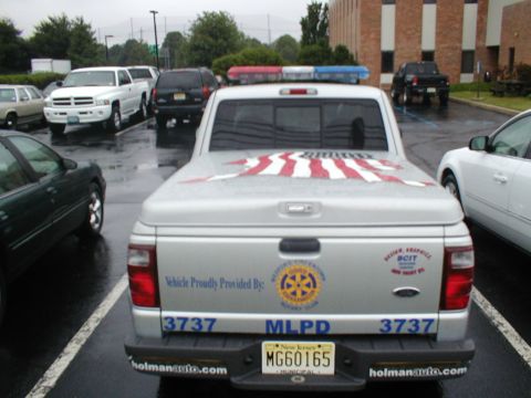 1979 Ford Bronco - Silver pickup truck with American flag decal on the bed cover and police lights on the roof is parked in a lot.
