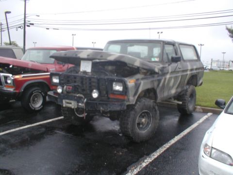 1979 Ford Bronco - A camouflaged Ford Bronco is parked with its hood up next to a red truck.