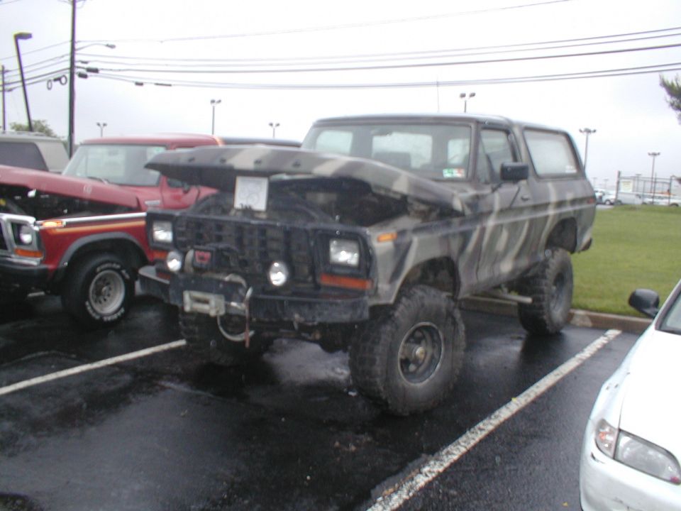 Camouflaged Ford Bronco with hood open, winch, and off-road tires parked on wet asphalt