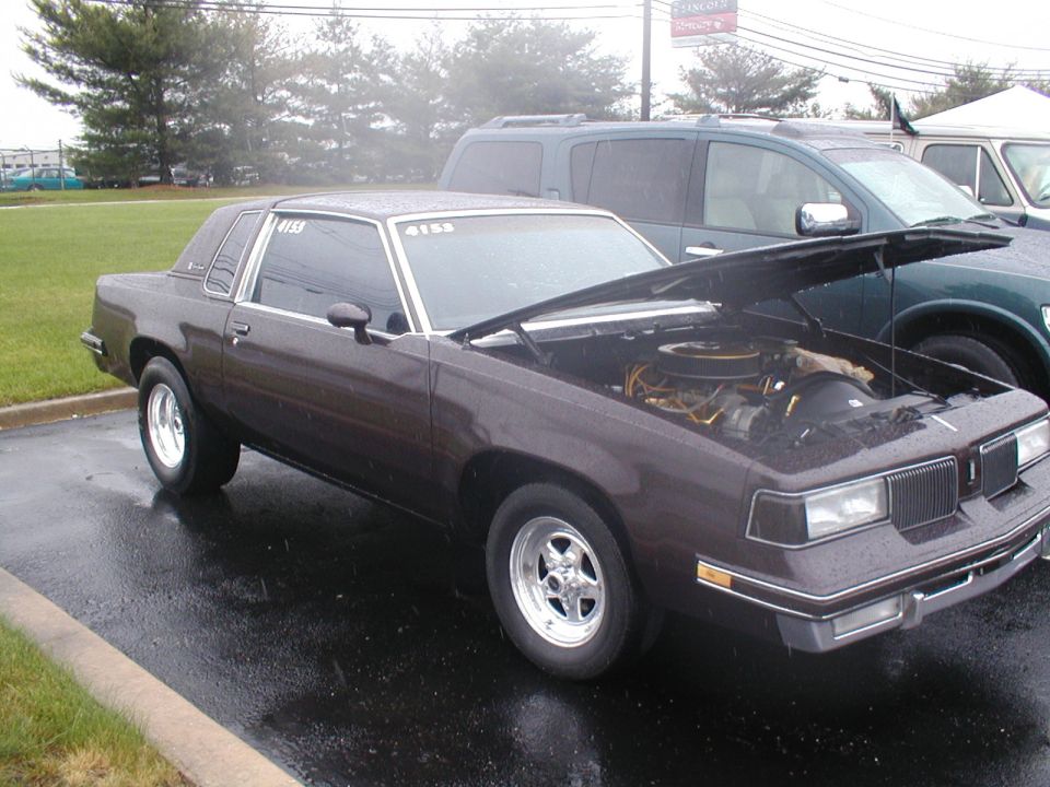 Dark brown 1981 Oldsmobile Cutlass coupe with hood open, parked next to a dark green SUV