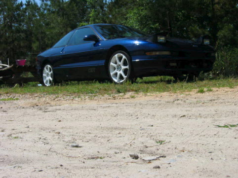 Dark navy-blue sport coupe with alloy wheels on a sandy dirt lot, front three-quarter view