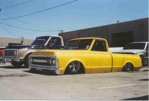 1990 Ford Bronco - bright yellow lowered Chevrolet pickup with chrome wheels sits in a lot beside a brown vintage pickup and a building.
