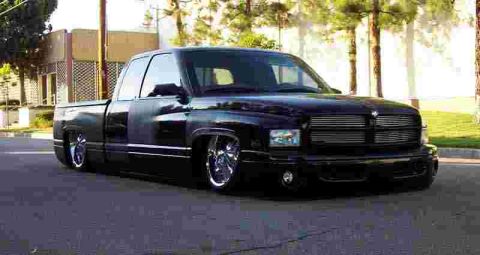 1990 Ford Bronco - glossy black lowered Dodge Ram extended-cab pickup with chrome wheels parked on pavement near trees.