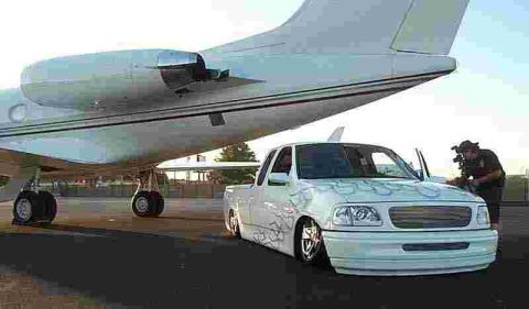 1990 Ford Bronco - white lowered Ford pickup with ghost flames sits on an airfield tarmac beside a parked white private jet, with a photographer nearby.
