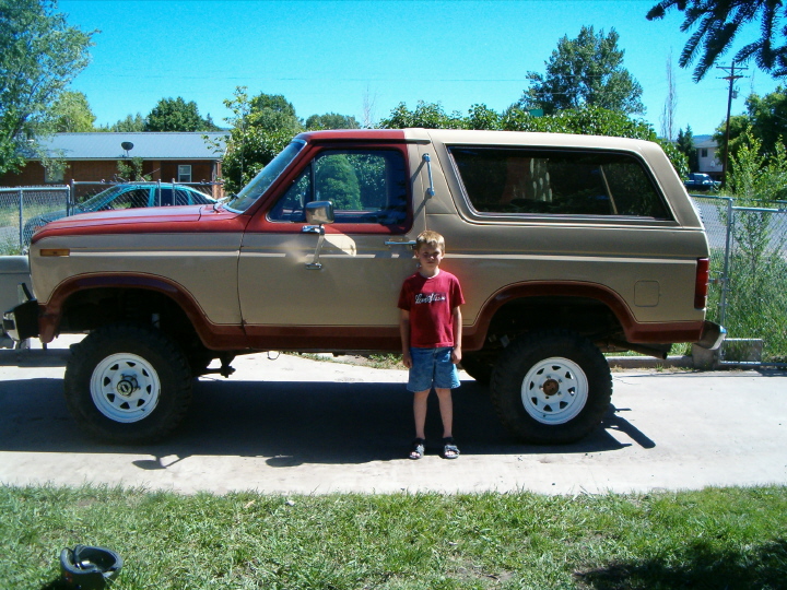 Lifted two-tone brown and tan Ford Bronco on white wheels with a boy standing beside it in a driveway