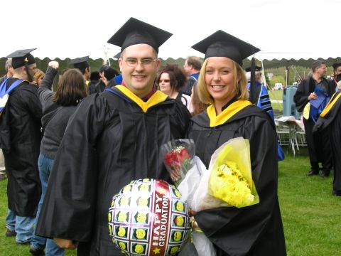 1998 Ford Taurus - Two graduates in black caps and gowns with yellow hoods pose on a grassy field, holding bouquets of red roses and yellow flowers and a "Hap…
