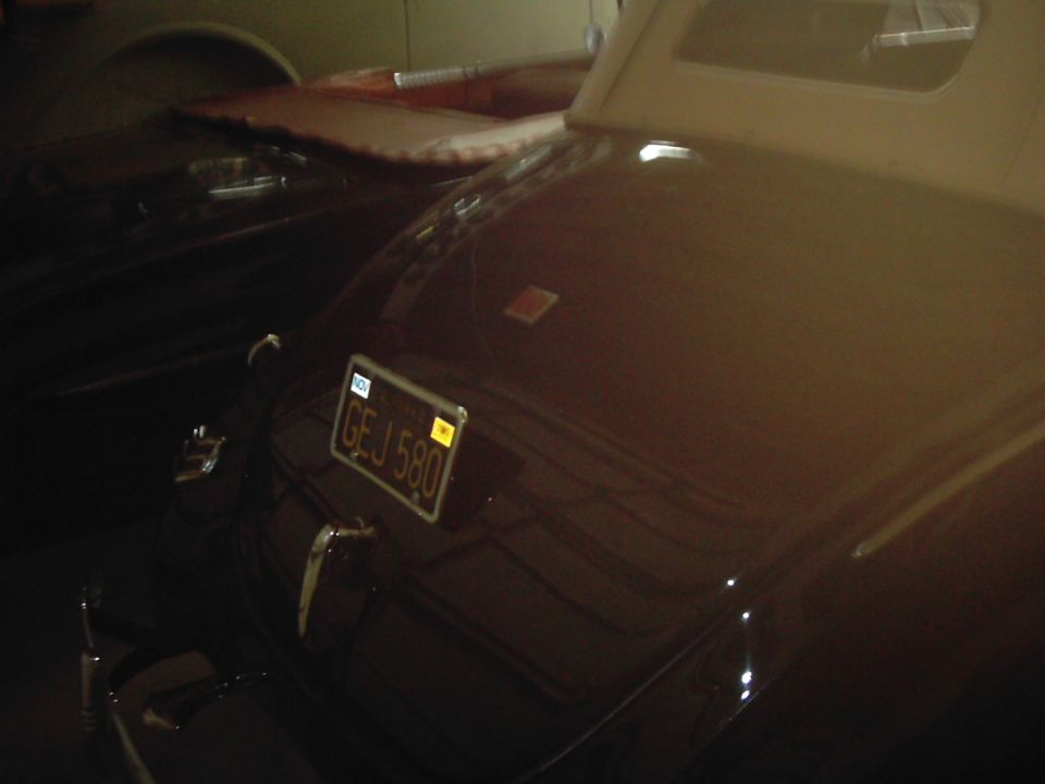Rear of a dark colored classic car showing license plate GEJ 580, with another car partially visible behind it