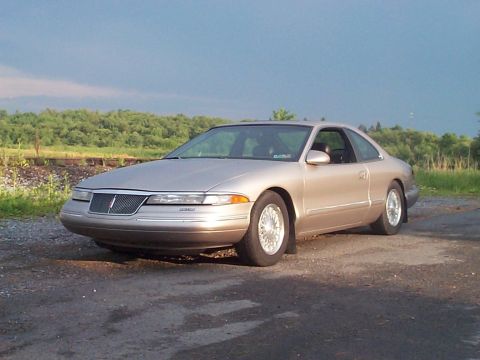 1900 Ford Wagon - A gold Lincoln Mark VIII coupe is parked outdoors on a gravel surface with a wooded area behind it.