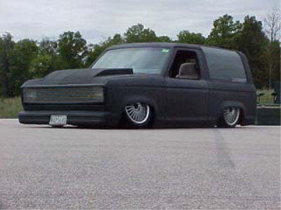 1990 Ford Bronco - flat-black slammed two-door SUV with cowl hood and custom wheels sits very low on a paved lot in front of trees.