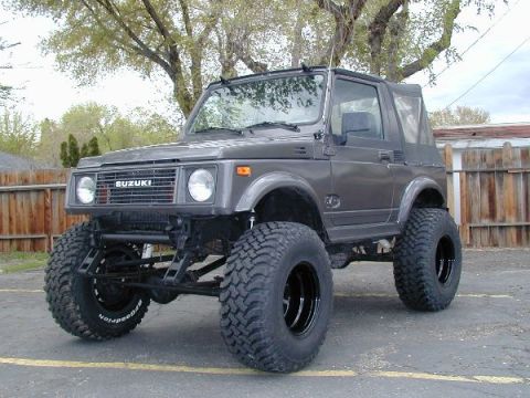 1990 Ford Bronco - gray lifted Suzuki Samurai 4x4 on oversized mud tires sits in a yard in front of a wooden fence and trees.