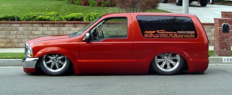 1990 Ford Bronco - red lowered two-door Ford Excursion SUV with custom wheels and graphics sits at a residential curb.