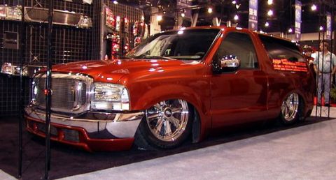 1990 Ford Bronco - glossy orange-red lowered Ford SUV with chrome wheels sits behind stanchions on an indoor show floor.