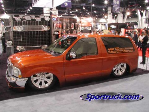 1990 Ford Bronco - bright orange lowered two-door Ford SUV with chrome wheels sits at an indoor trade show booth; a 'Sportruck.com' watermark is overlaid.