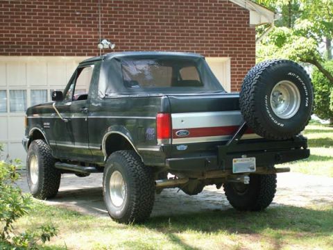 1988 Ford Bronco - Three-quarter rear view of a lifted black full-size Ford Bronco parked on grass in front of a brick garage, with a black soft top over the…