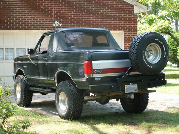 Lifted black Ford Bronco soft top rear 3/4 with swing-out BFG spare by brick garage