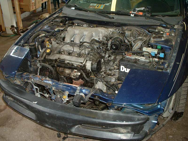 Car engine bay with hood open, showing engine, battery, and front end damage