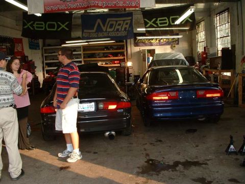 1994 Ford Probe - Two coupes are parked in a garage with various banners and shelves in the background.