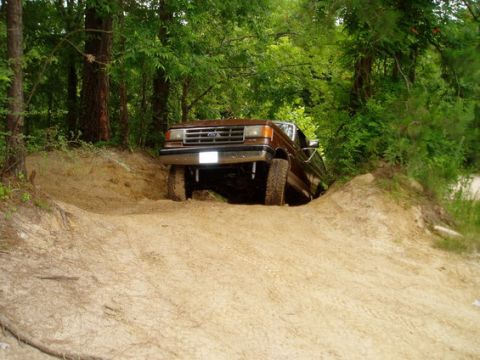 1987 Ford Bronco - A brown 1987 Ford Bronco navigates a challenging off-road dirt track surrounded by trees.