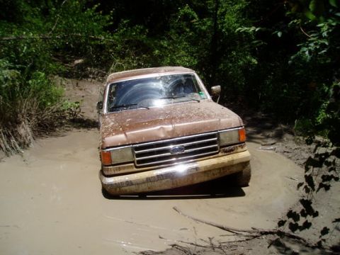 1987 Ford Bronco - This Ford truck is navigating a challenging muddy terrain, with its front wheels deep in the mire.