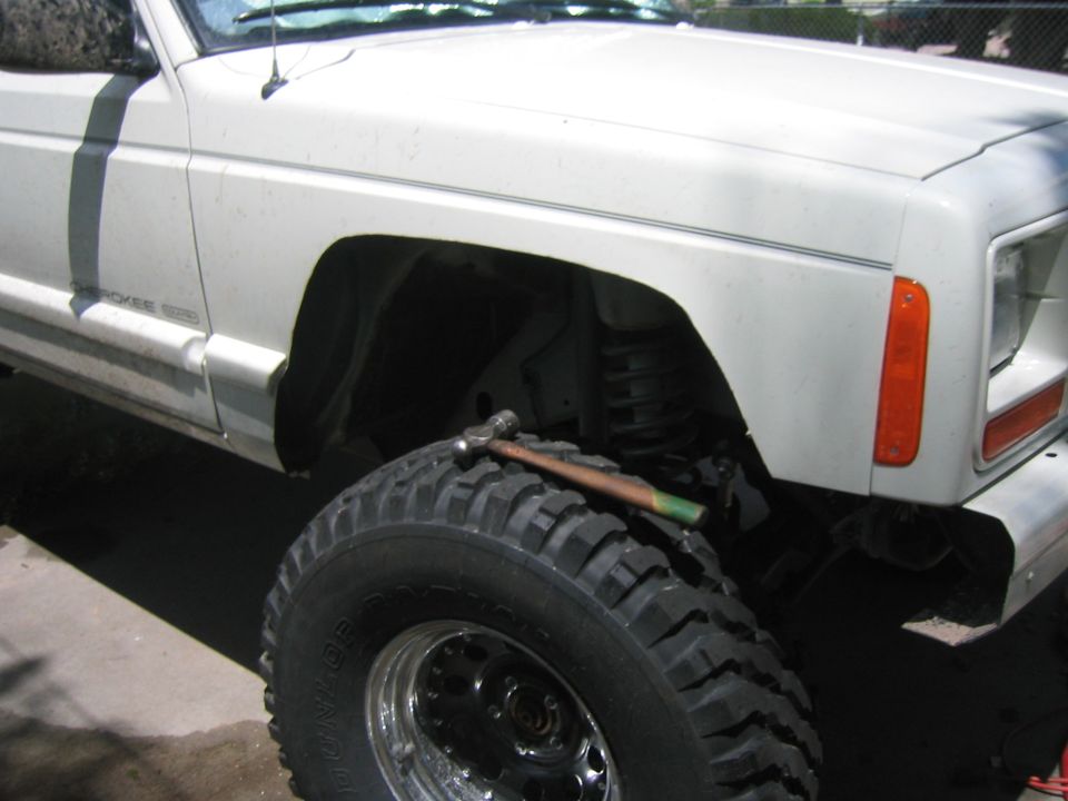 Trimmed front fender of a white Jeep Cherokee showing coil spring and mud tire on chrome wheel