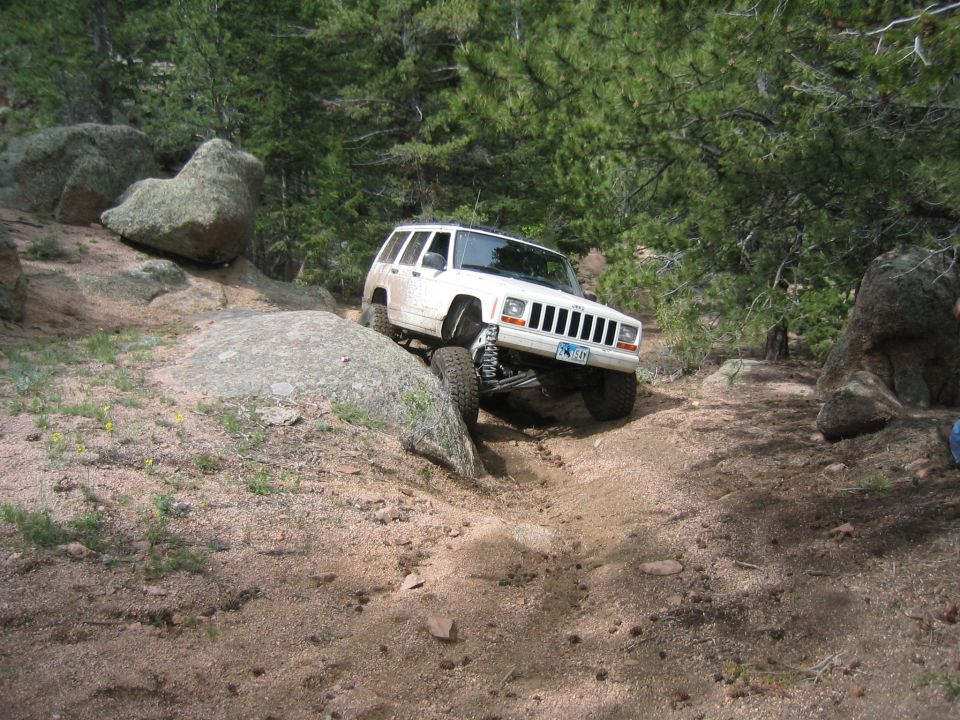 White lifted Jeep Cherokee XJ descending a rocky sandy trail in pine forest