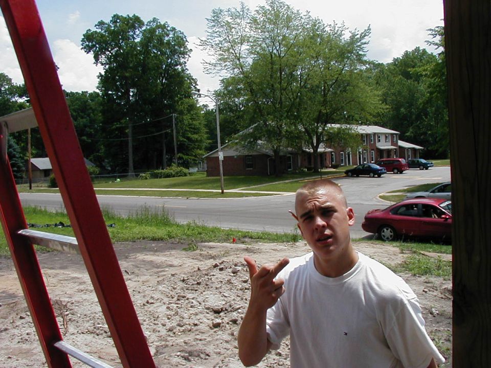 Man in white t-shirt gestures with fingers in front of apartment buildings and parked cars