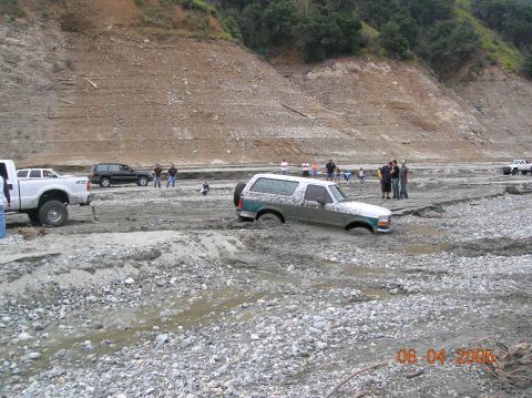 1985 Ford Bronco - Multiple vehicles are stuck in a muddy riverbed with people observing.