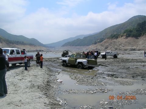 1985 Ford Bronco - Several off-road vehicles are stuck in mud and rocks in a wide, dry riverbed with hills in the background.