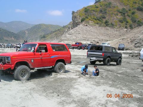 1985 Ford Bronco - Two Ford Broncos are seen in a dusty, off-road environment, with the red one towing the black one.