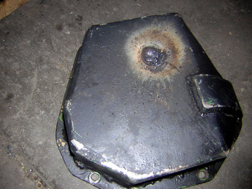 Black oil pan or differential cover with a burned scorched spot near a drain plug on a concrete floor