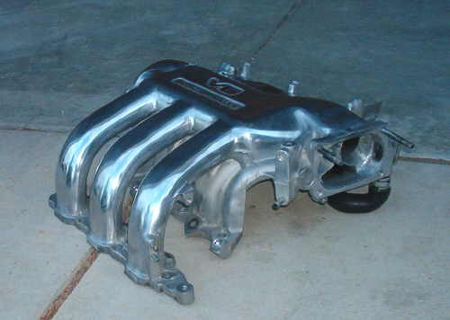 Polished aluminum intake manifold with four runners, resting on a concrete floor