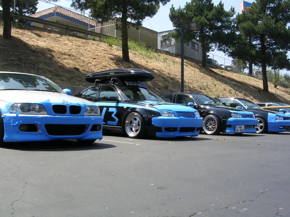 Several cars parked in a row, all with blue tape on their front bumpers and some with roof racks