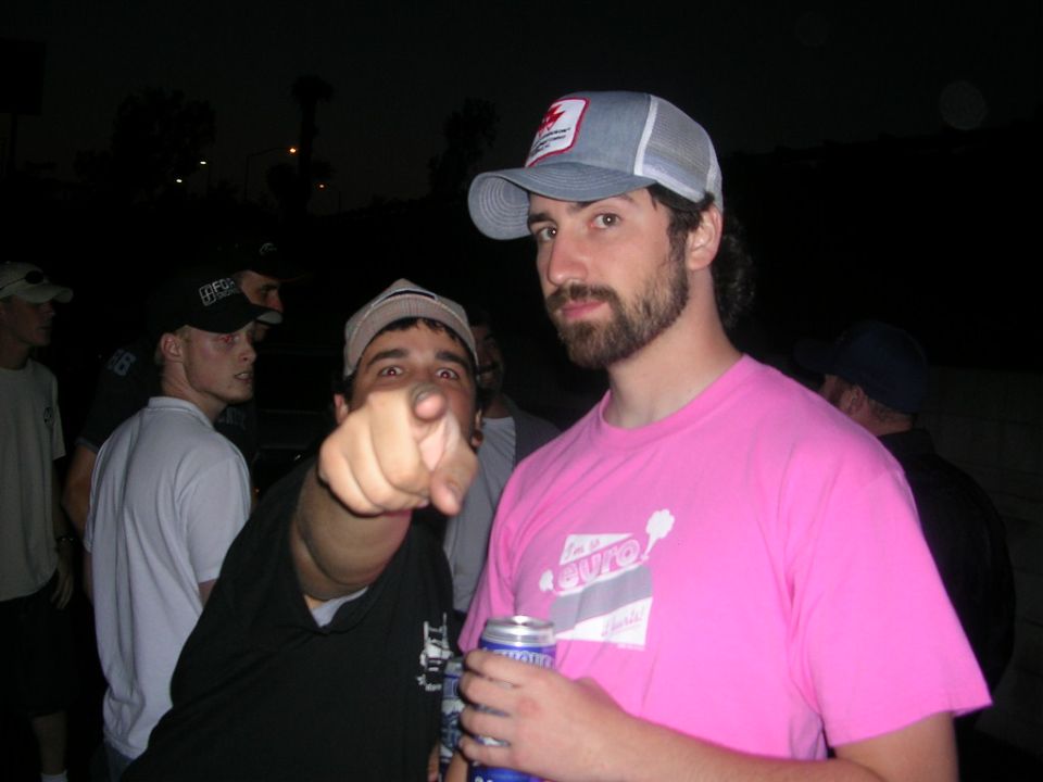 Guy in pink shirt with beard and cap, another guy pointing at camera, others in background at night