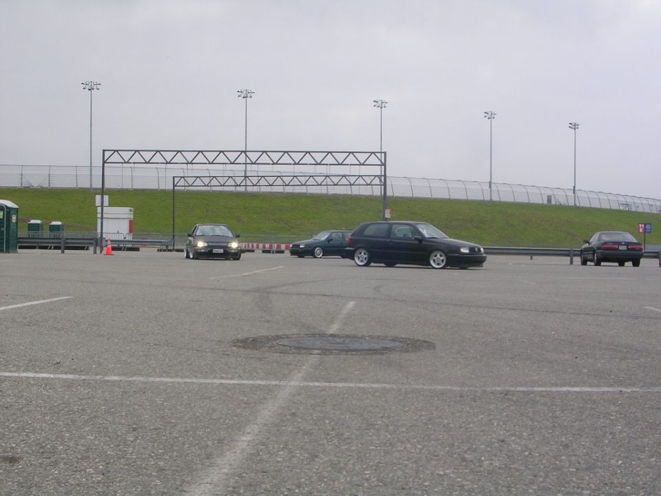 Several cars are parked and driving in a lot with overhead lights and a fence in the background