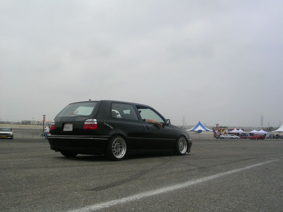 Black Volkswagen Golf GTI on a track, driver's arm out window. Tents and other cars in background