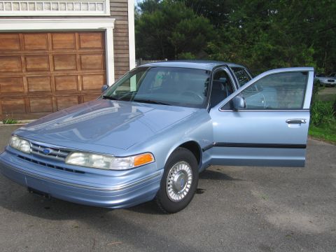1991 Ford LTD Crown Victoria - A light blue sedan is parked in front of a wooden garage door, with the driver's side door ajar.