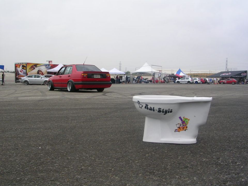 Toilet with 'Rat-Stylz' sticker and cartoon graphics, parked on asphalt with cars and tents in background