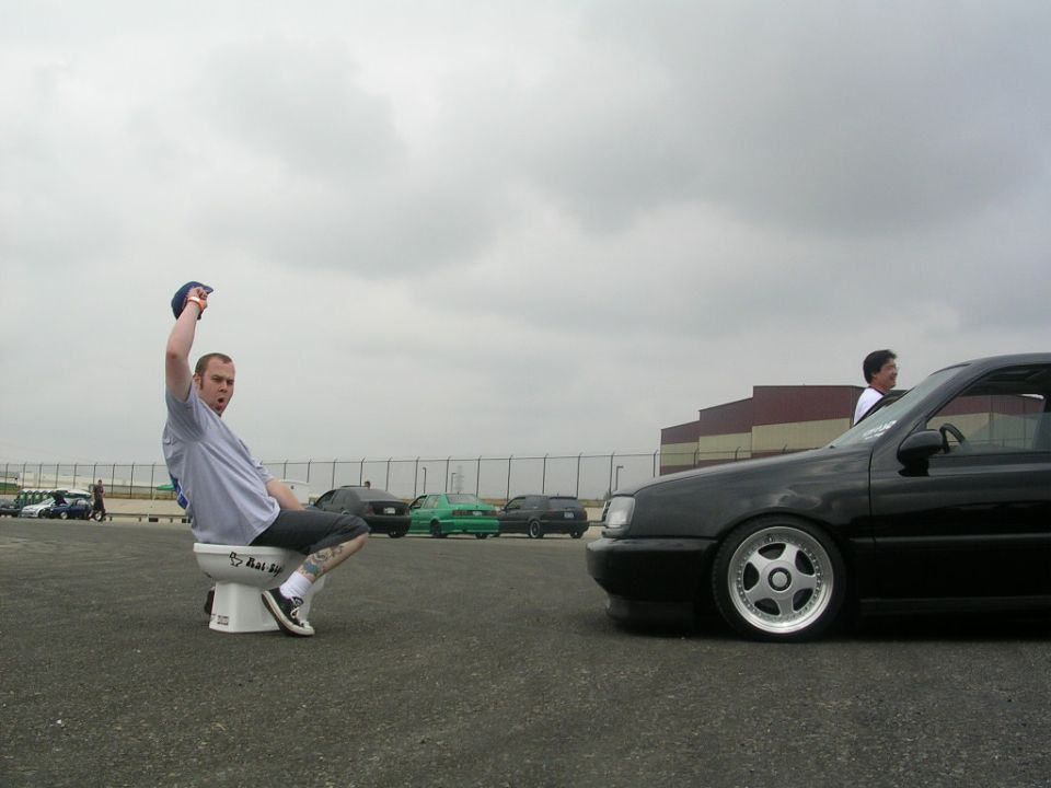 guy on toilet next to black car, other cars in background