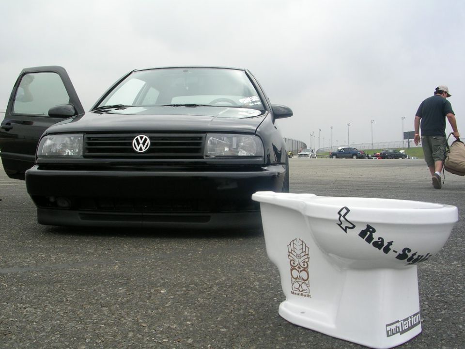 Black Volkswagen with driver's door open, parked on asphalt with a toilet in the foreground