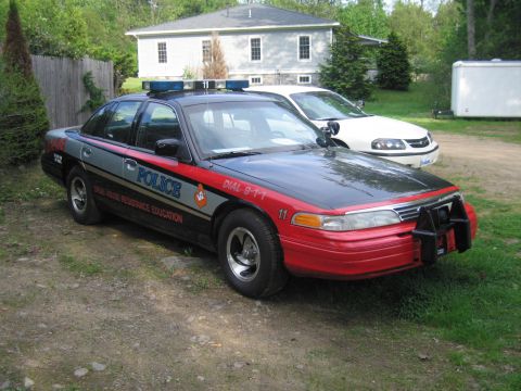 1991 Ford LTD Crown Victoria - A police car with a black and red paint scheme is parked in front of a house, with another white car behind it.